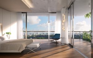 A look inside a master bedroom at Monad Terraces, room includes a large bed with white linen, a modern blue chair, and ceiling to floor glass windows overlooking Biscayne Bay